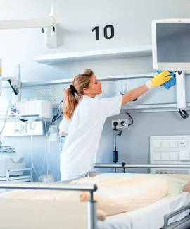 Eine Frau in weißer Kleidung reinigt die Ablage oberhalb eines Krankenhausbettes. Im Hintergrund sind Bildschirme und medizinische Technik zu erkennen.